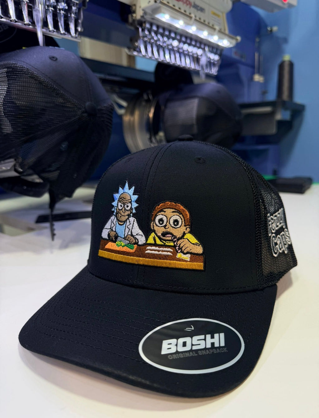 Black cap with cartoon characters and 'Boshi' logo on a white surface with embroidery machine in the background